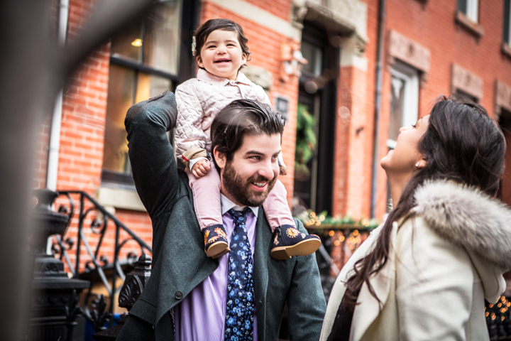 A man carries a laughing child on his shoulders beside a woman on a city sidewalk.