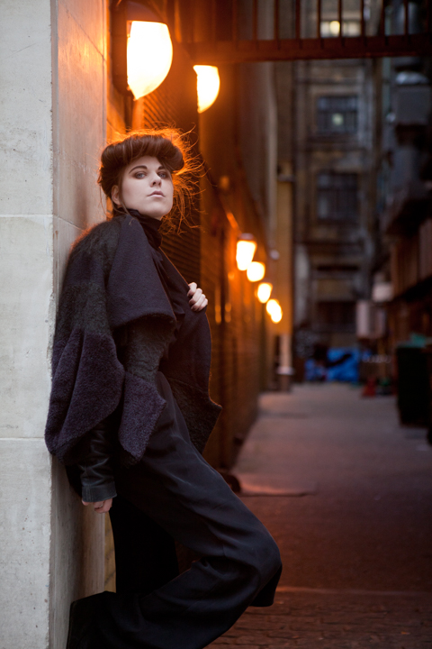 A woman in dark clothing leans beside a warmly lit doorway.