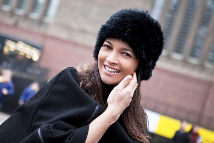 A smiling woman wearing a black fur hat on a city street.