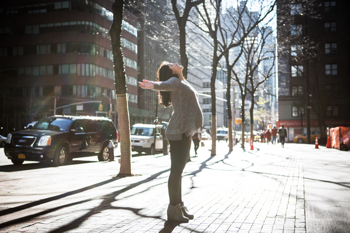 A person stretches their arms beside a tree on a sunny city sidewalk.