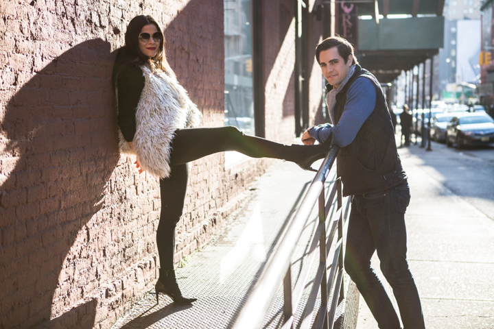 Two adults lean against a wall and railing on a sunny city street.