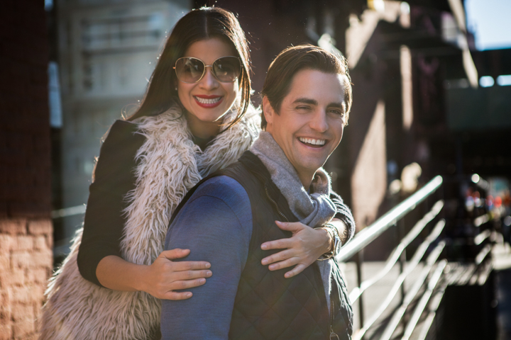 A smiling man carries a woman on his back beside a brick building.