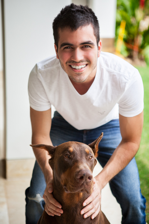A smiling man in a white shirt crouches behind a brown dog.