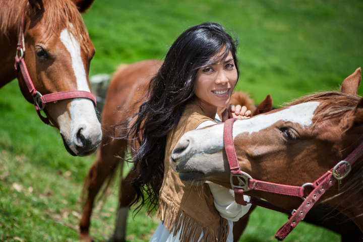 A woman stands between two brown horses in a green pasture.