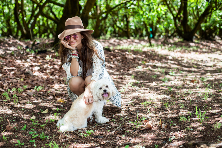 A woman in a hat crouches beside a small white dog under trees.