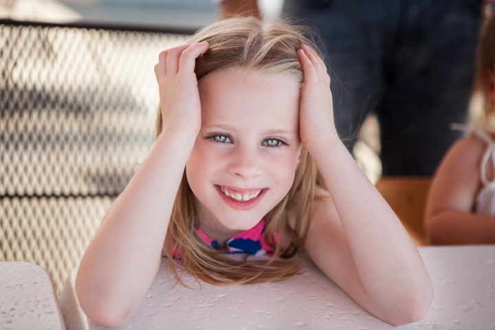 A smiling child rests their chin on their hands beside an adult indoors.