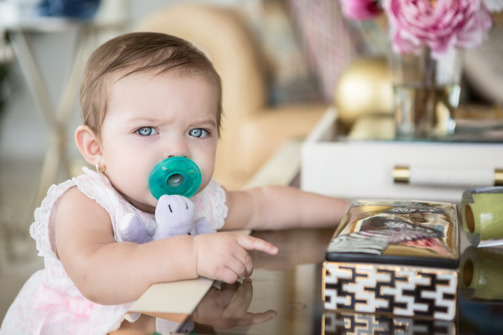 A baby with a green pacifier leans against a table indoors.