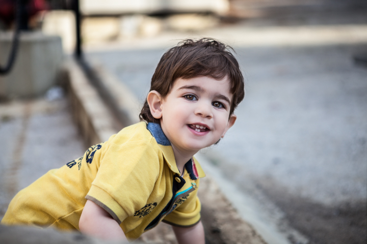 A smiling child in a yellow shirt leans forward outdoors.