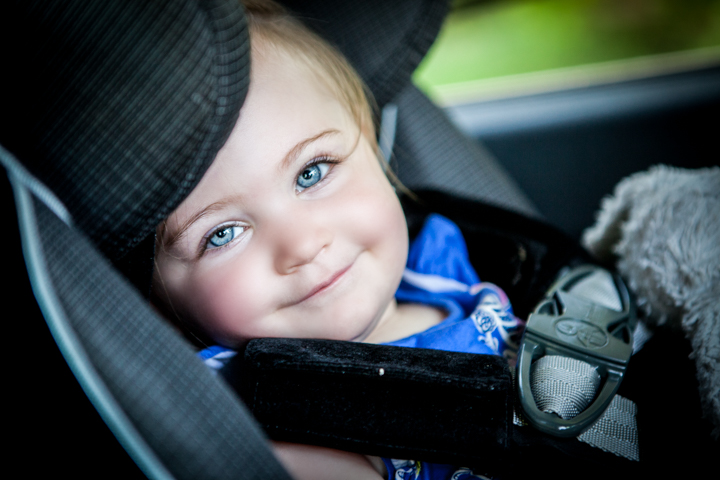A child wearing a dark hat smiles from a car seat.