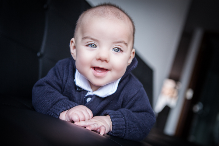 A smiling baby in a dark cardigan rests their hands on a surface.