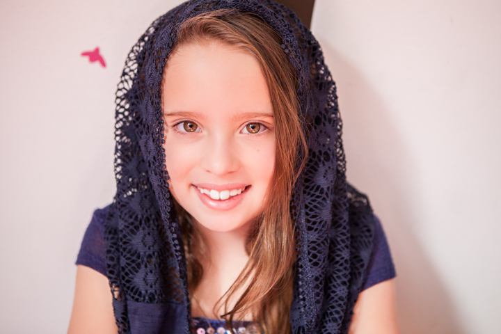A smiling child wears a dark lace scarf over their hair.