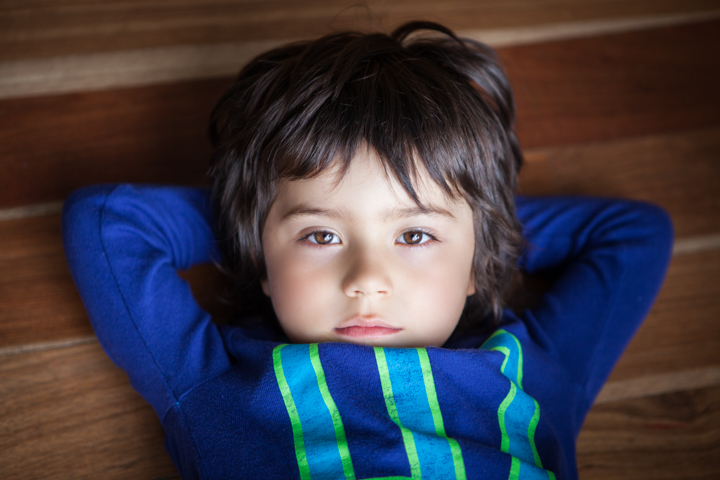A child lies on a wooden floor with both hands behind their head.