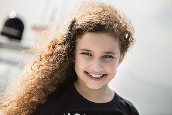 A smiling child with curly hair stands outdoors in a black top.