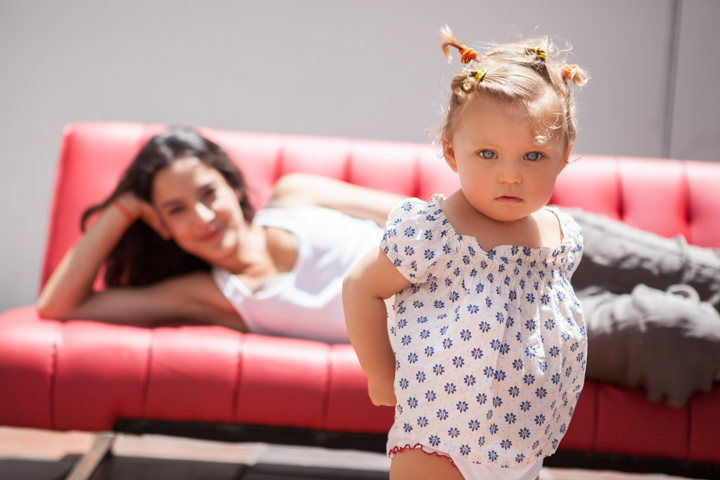 A toddler stands in front of a red sofa where a woman reclines.