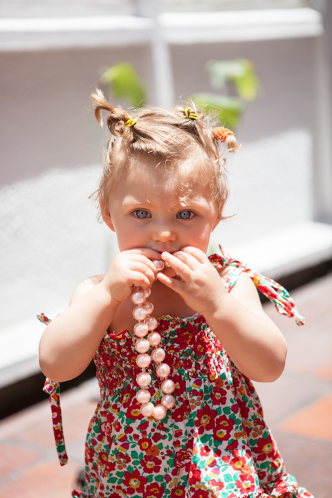 A toddler in a floral dress holds their hands near their mouth outdoors.