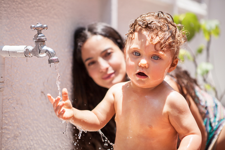 A toddler plays beside an outdoor tap while a woman watches.