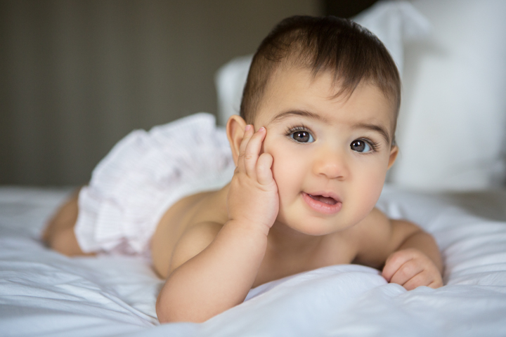 A baby rests on a white bed with one hand against their cheek.