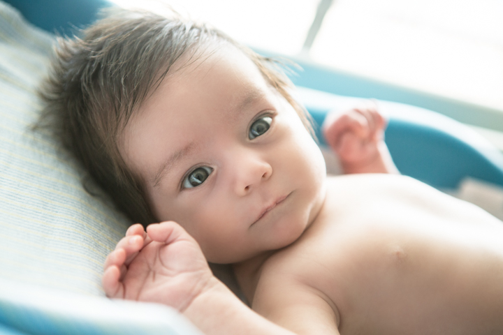 A baby lies in a bath with both hands lifted beside their face.