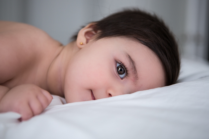 A baby lies on white bedding and looks toward the camera.