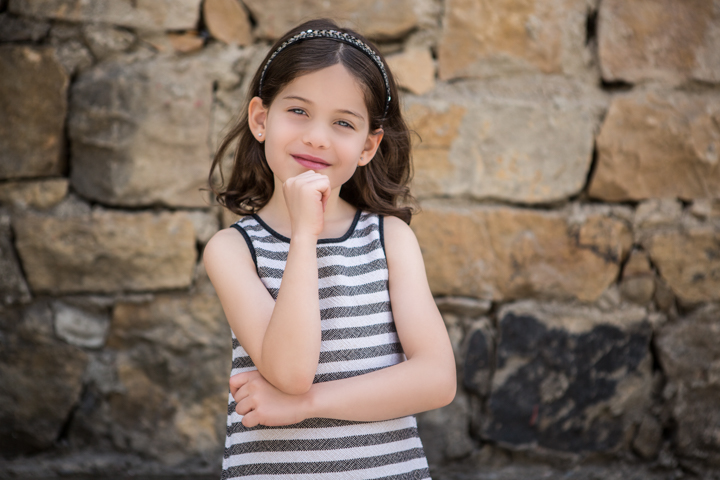 A child in a striped dress rests a finger on their chin beside a stone wall.
