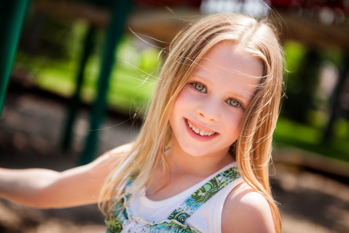 A smiling child with long blonde hair sits outdoors in sunlight.