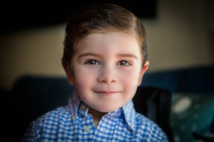 A smiling child in a blue checked shirt looks toward the camera indoors.