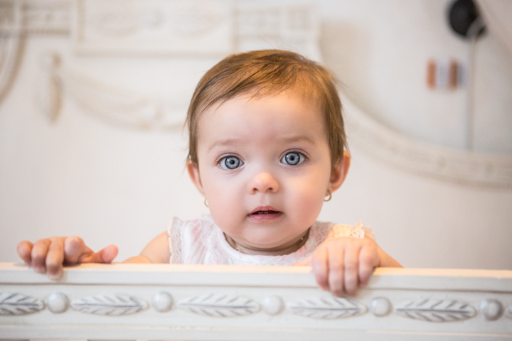 A baby holds the edge of a pale crib and looks toward the camera.