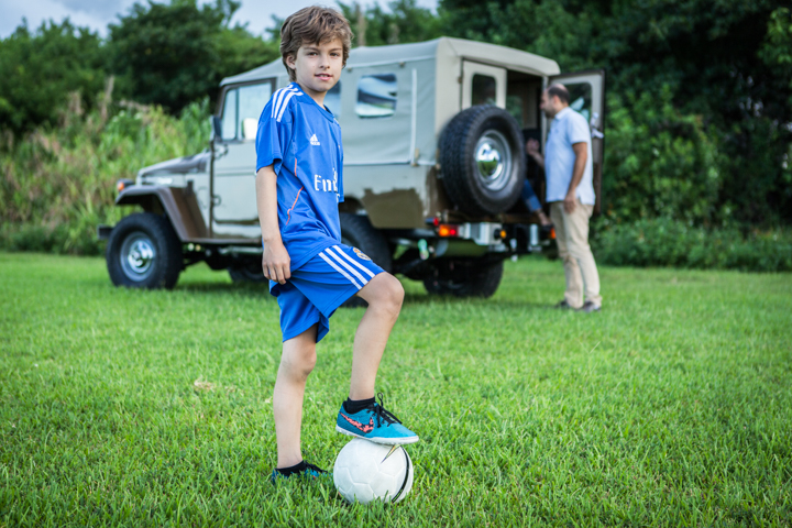 A child in a blue soccer kit rests a foot on a ball in a grassy field.