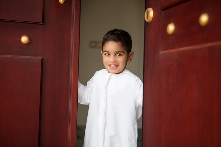 A smiling child in white stands between two dark red doors.