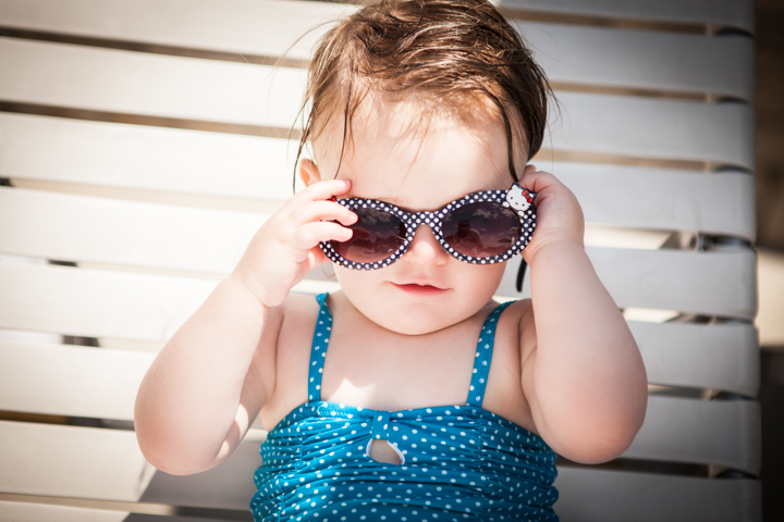 A toddler adjusts oversized sunglasses while sitting on a white outdoor chair.