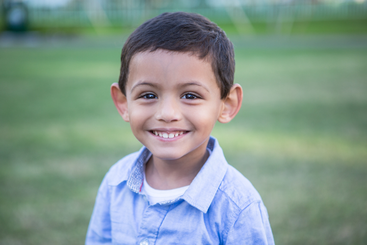 A smiling child in a pale blue shirt stands in a green park.