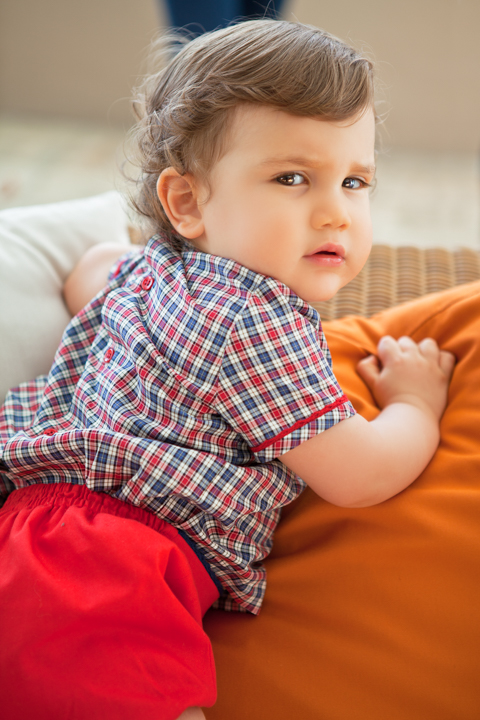 A young child in a checked shirt and red shorts leans on an orange cushion.