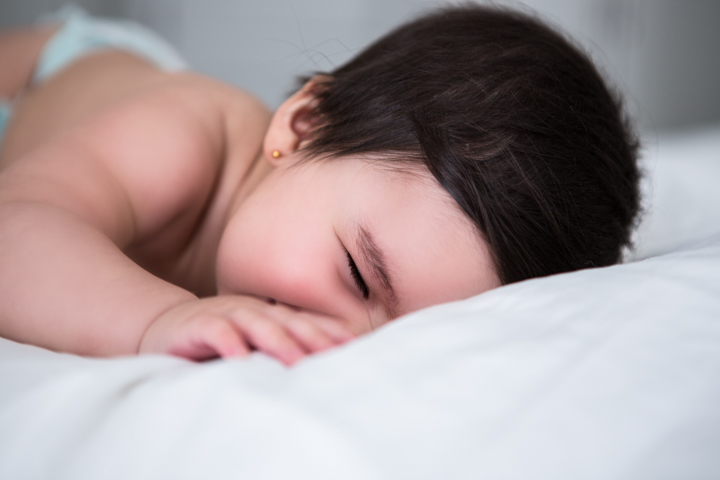 A baby sleeps on white bedding with a hand near their face.