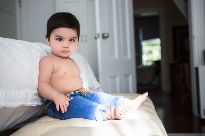 A toddler in blue jeans sits against white cushions.