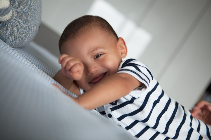 A laughing baby in a striped top rests against a pale cushion.