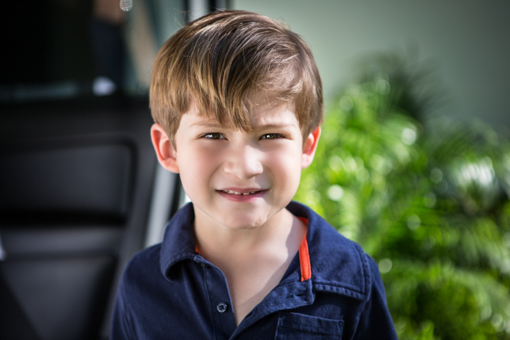 A smiling child in a dark polo shirt stands outdoors.