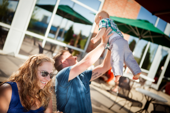 A smiling woman lifts a small child into the air outdoors.