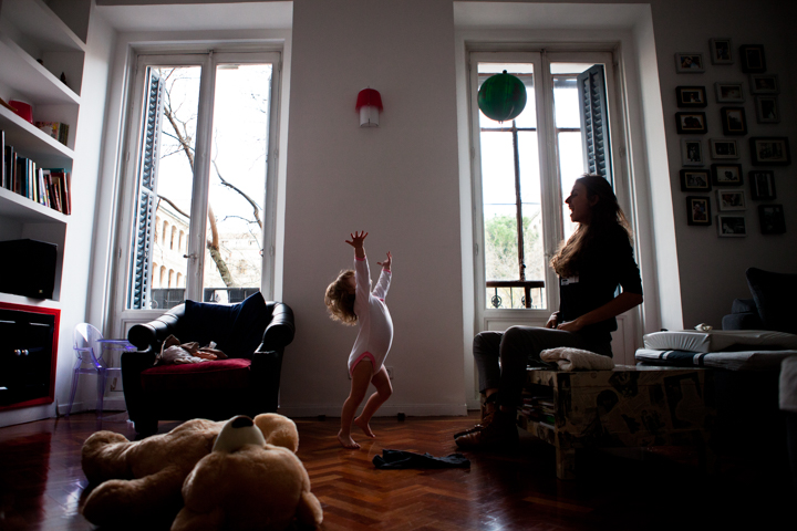 A child raises both arms in a sunlit living room while a woman watches.