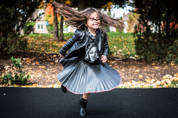 A child twirls a pleated skirt outdoors with trees behind.