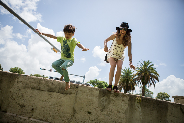 A child and woman balance with arms outstretched on a low outdoor wall.