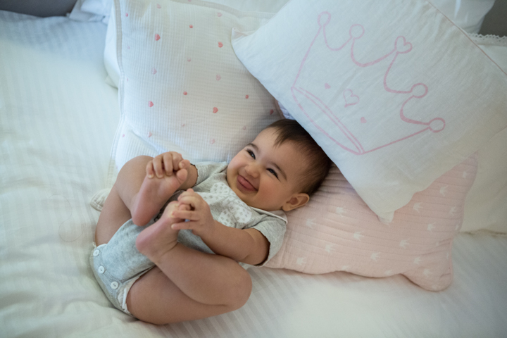 A smiling baby lies beneath pale pillows and holds their feet.