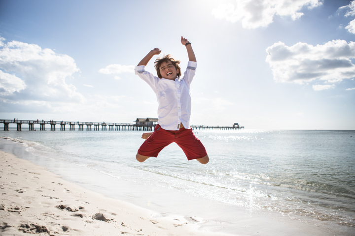 A child in red shorts jumps on a sunny beach with arms raised.