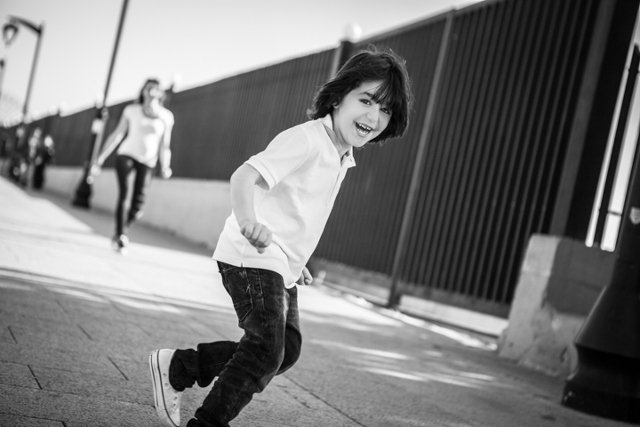A smiling child runs beside a dark fence in black and white.