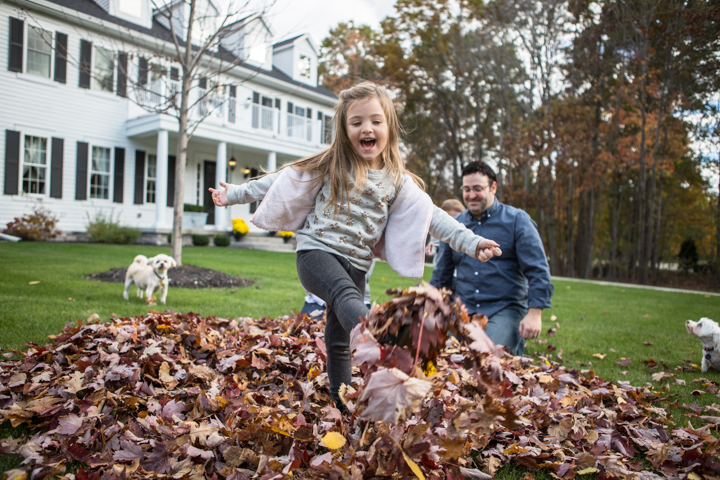 A child runs through a pile of autumn leaves while an adult watches.