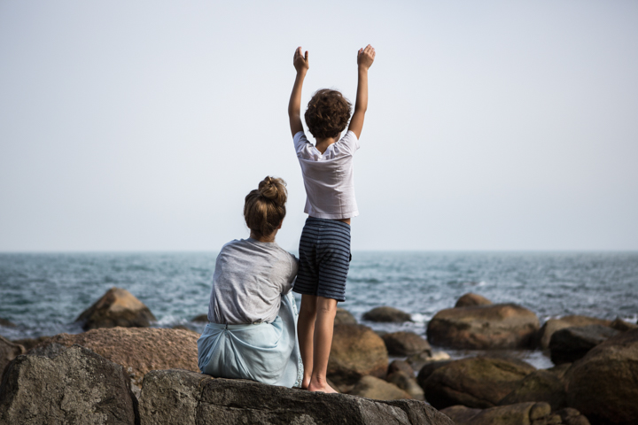 Two children stand on coastal rocks, one with both arms raised toward the sea.