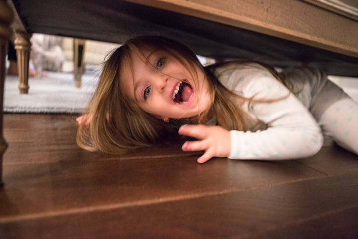 A laughing child peers from beneath furniture on a wooden floor.