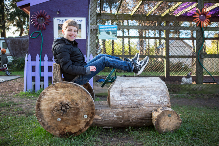A child sits on a wooden log beside colorful garden decorations.