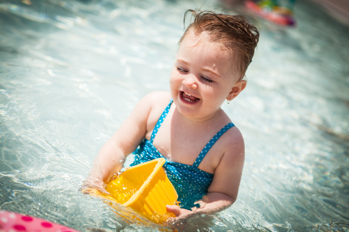 A laughing toddler plays in a swimming pool with a yellow bucket.