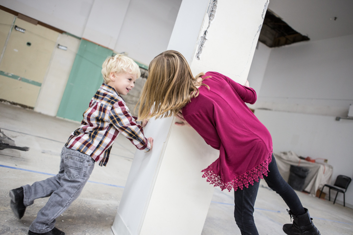 Two children lean toward each other beside a white wall indoors.