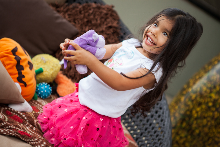 A smiling child in a pink skirt holds a stuffed toy beside a pile of toys.